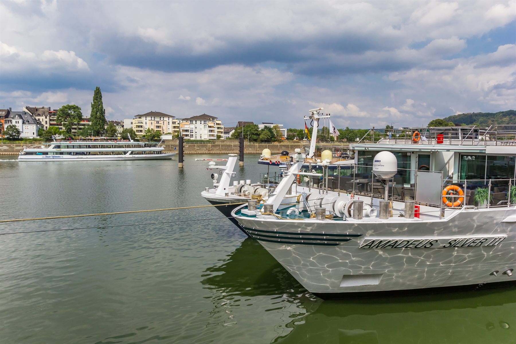 Bows of two moored river cruise ships, including Amadeus Silver III, with a day-trip boat passing